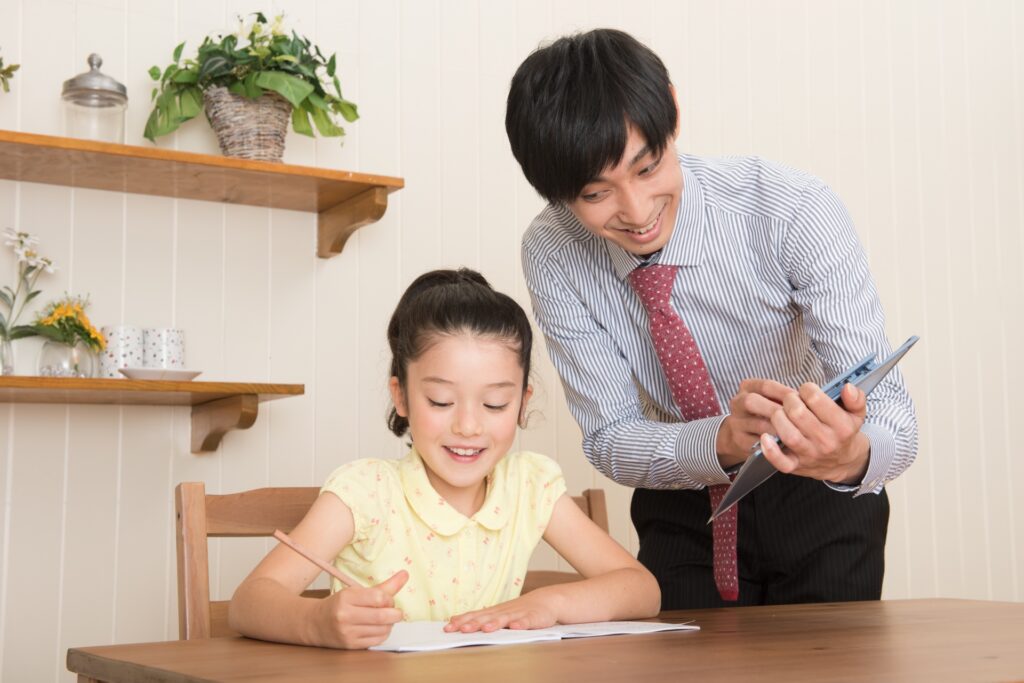 紙に書きながら勉強をしている笑顔の女の子と指導しているしましまのワイシャツを着た男性教員